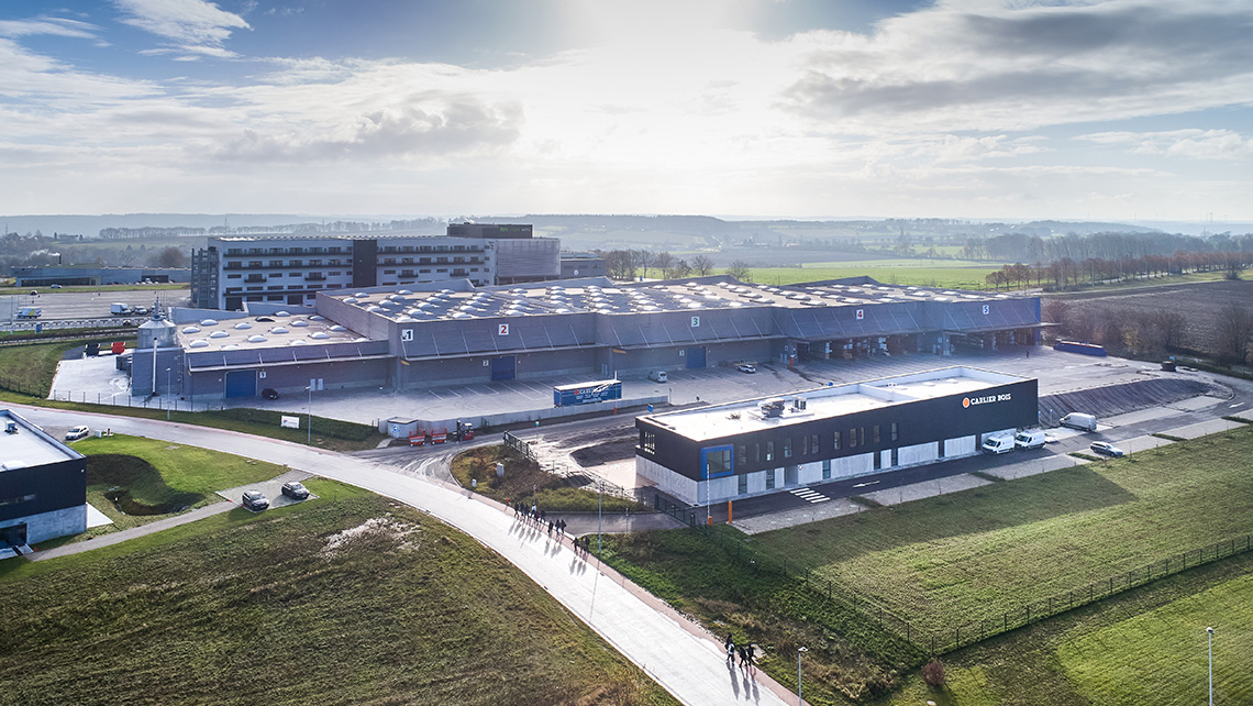 Une vue aérienne d'un grand complexe industriel ou logistique. Le site comprend plusieurs grands bâtiments avec des toits équipés de lucarnes et de panneaux solaires. Il y a des zones de stationnement pour les véhicules et les camions, ainsi que des espac